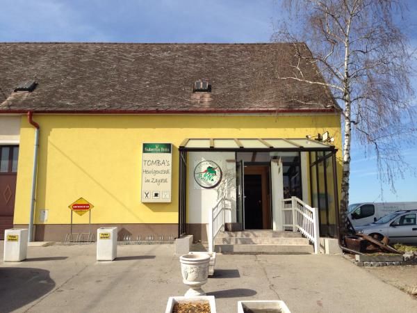 Žltá budova s nápisom "Tomba's Heurigenschenke". Vchod so schodiskom a sklenenou strechou.