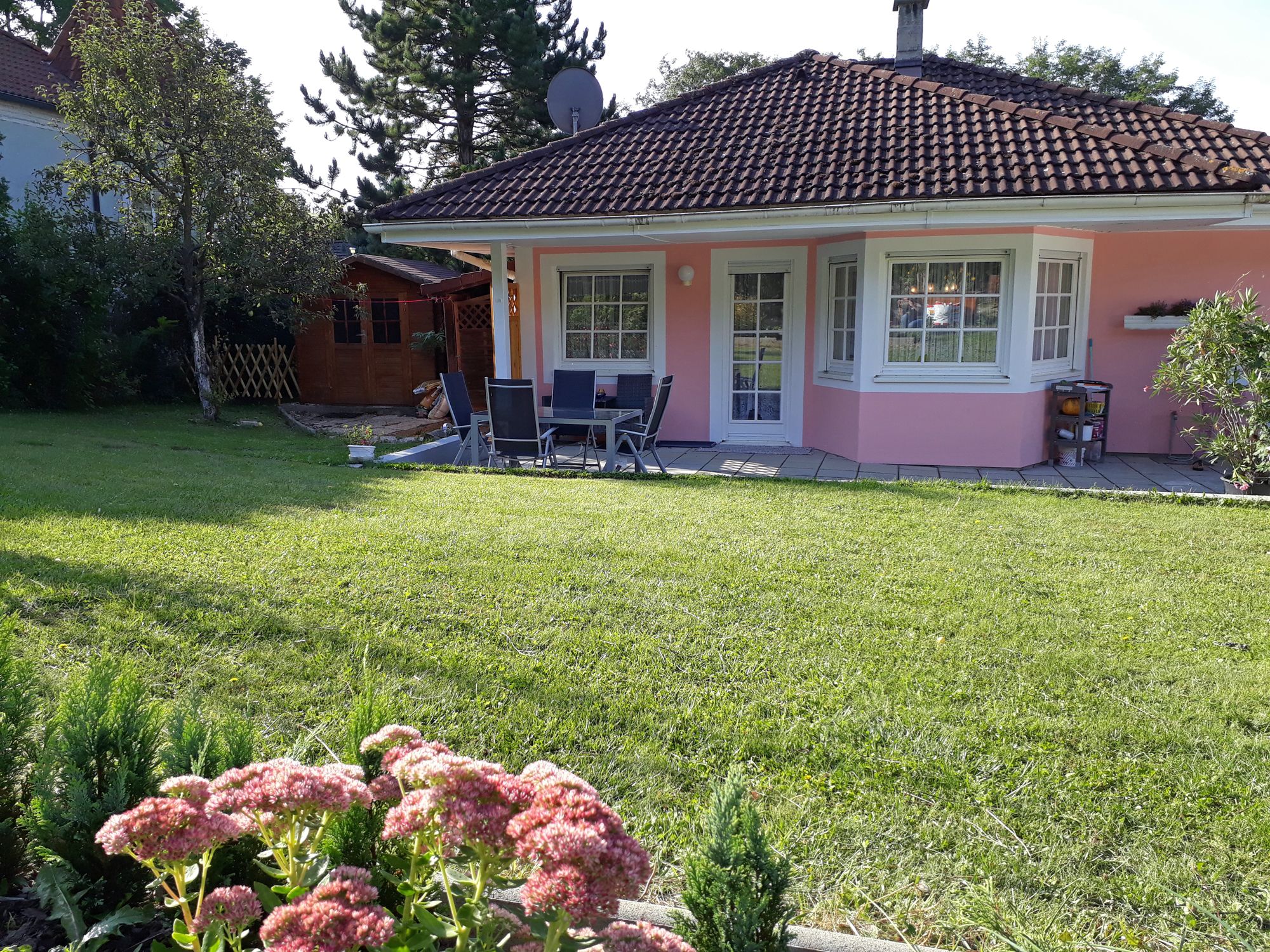 Ružový dom so záhradou, terasou a záhradným nábytkom, obklopený stromami a kvetmi.