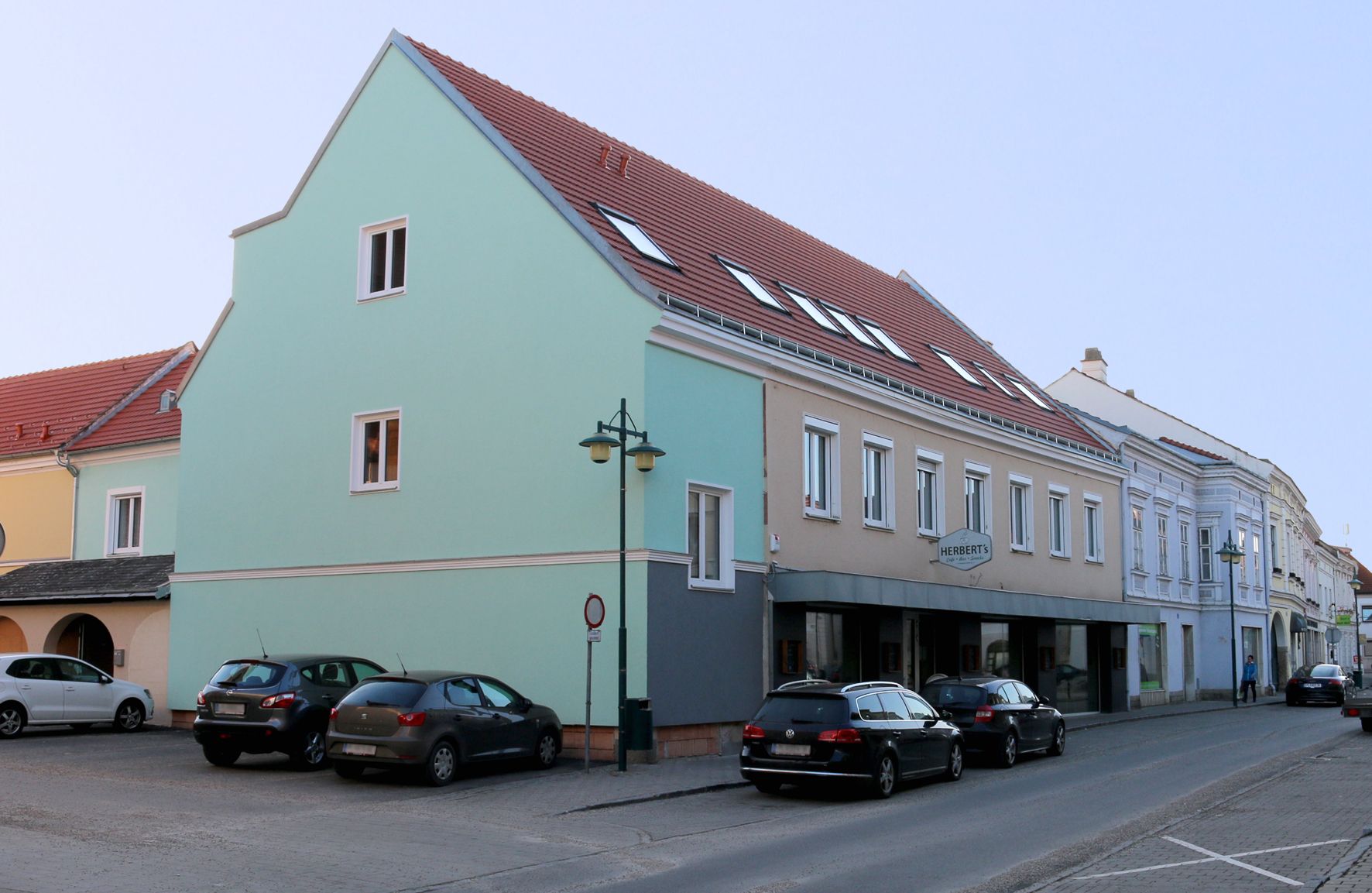 Scéna z ulice Traismauer s viacposchodovou budovou a zaparkovanými autami.