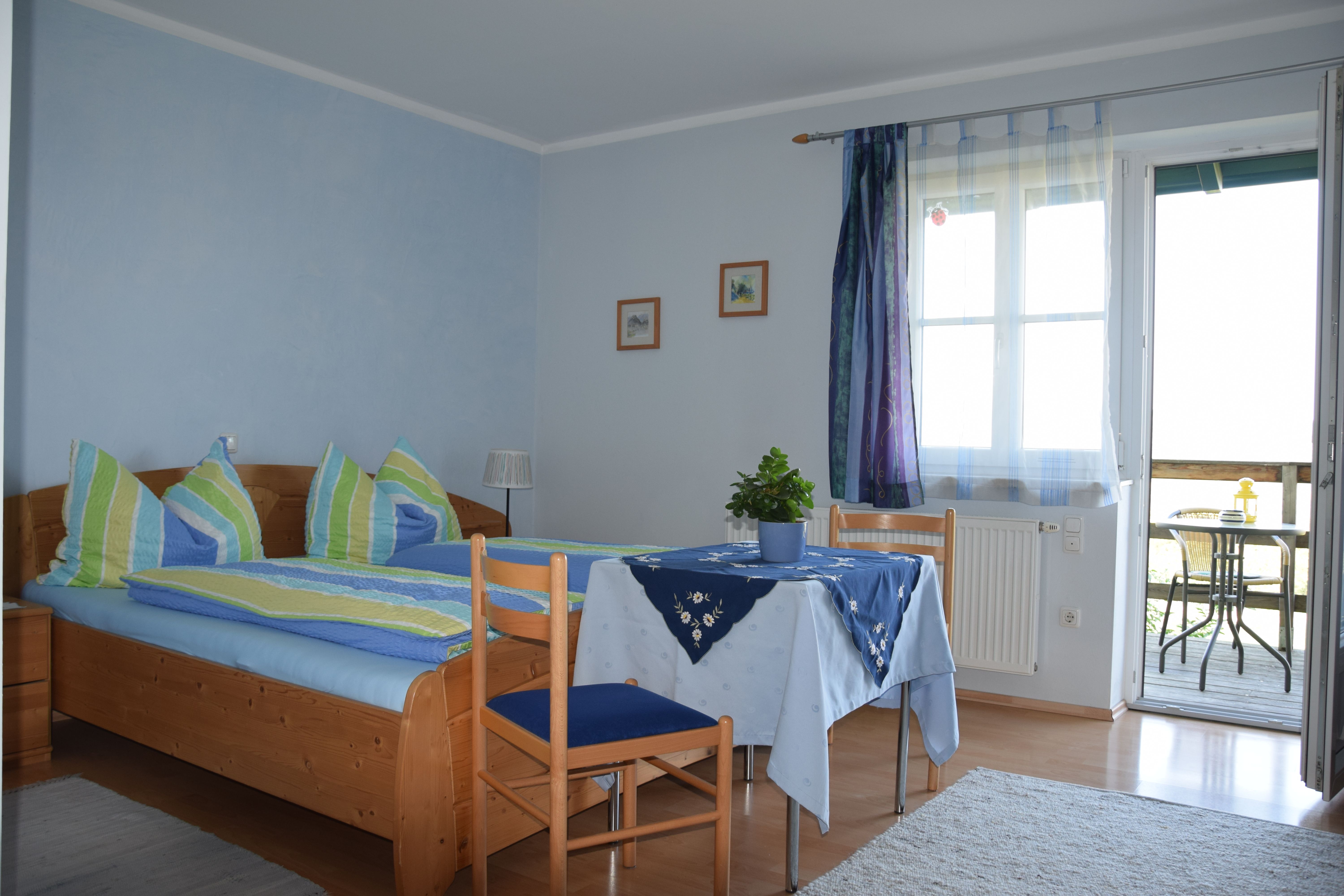 Útulná izba pre hostí s manželskou posteľou, stolom a stoličkami, modrými závesmi a prístupom na balkón s posedením.