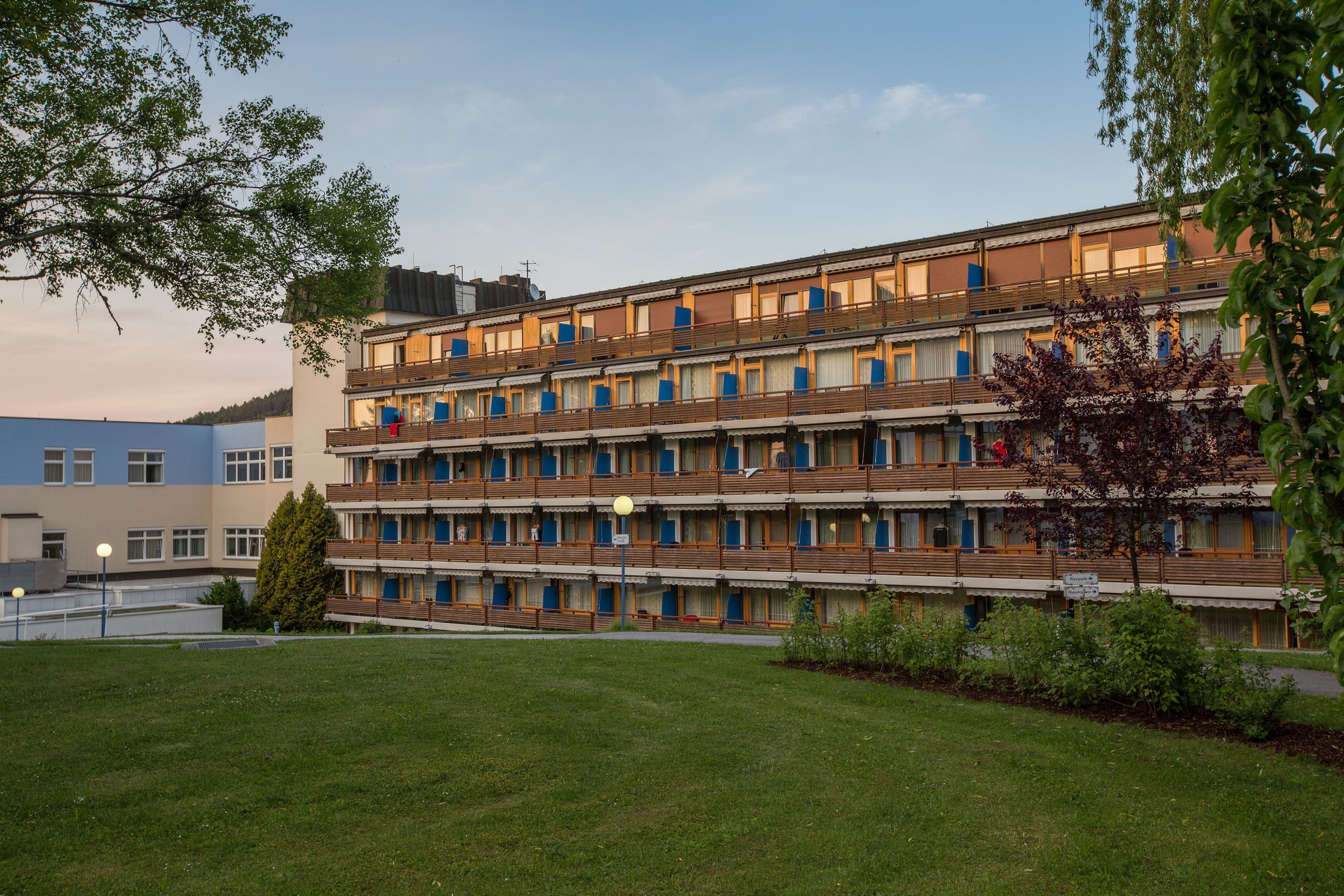 Viacposchodová budova s balkónmi a modrými markízami, obklopená stromami a trávnikmi.