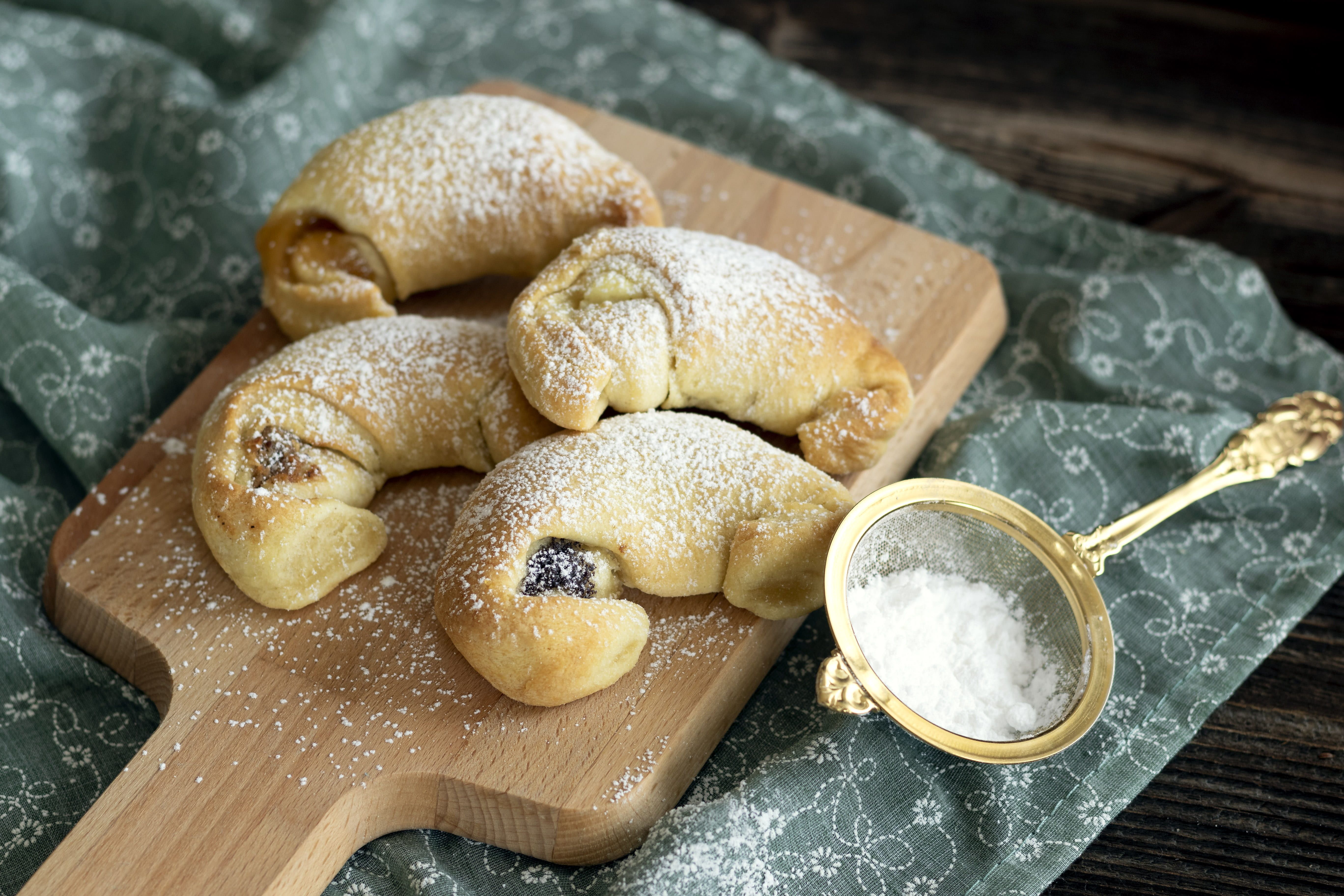 Štyri krémové croissanty na drevenej doske posypanej práškovým cukrom, vedľa sita s práškovým cukrom.