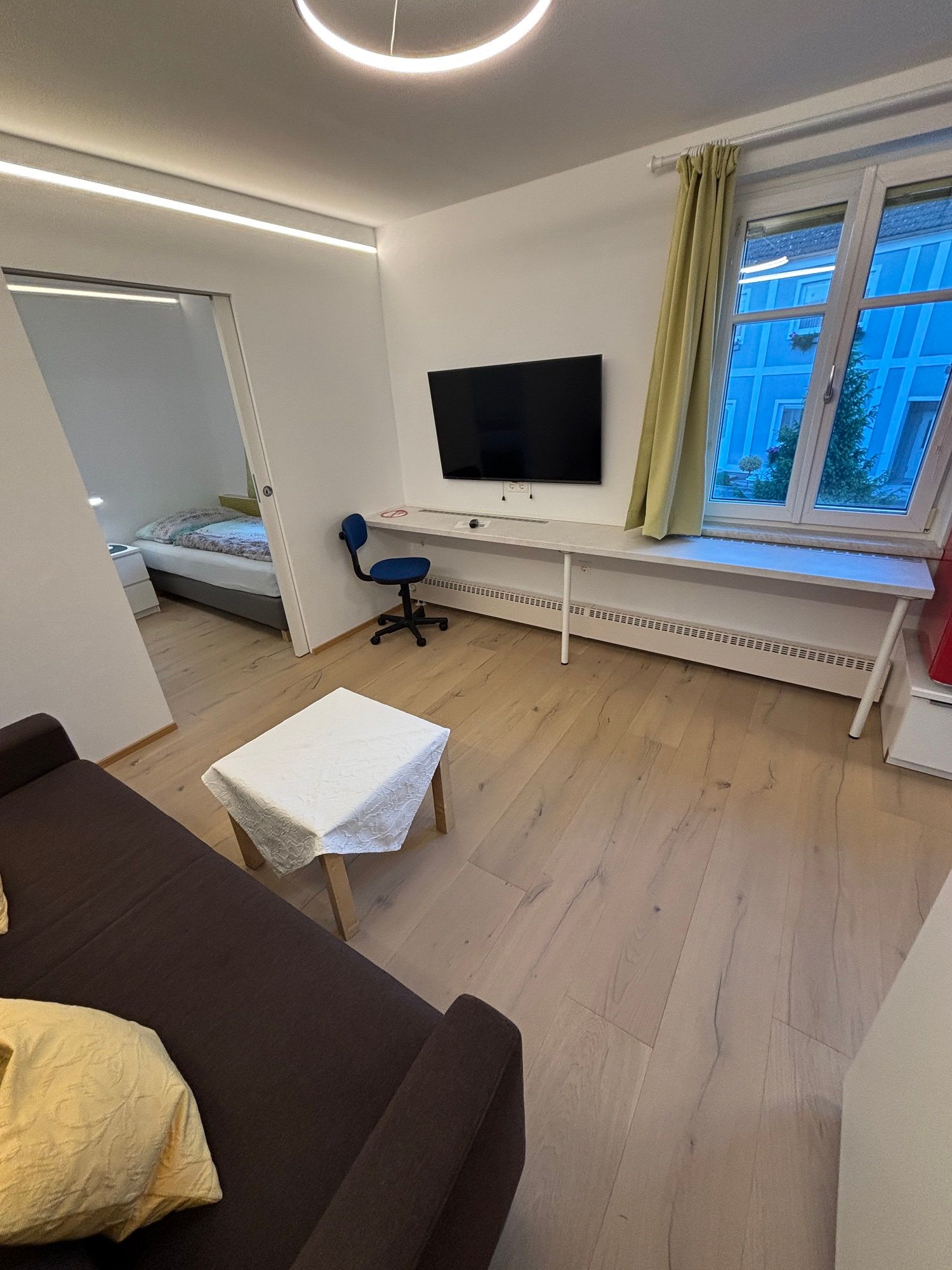 Moderná obývacia izba s hnedou pohovkou, stolíkom, TV na stene, písacím stolom a stoličkou. Okno s výhľadom na modrú budovu.