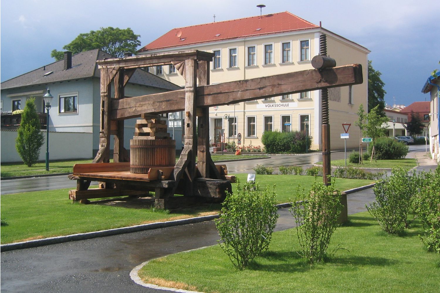 Historický lis pred budovou školy v zelenom prostredí.