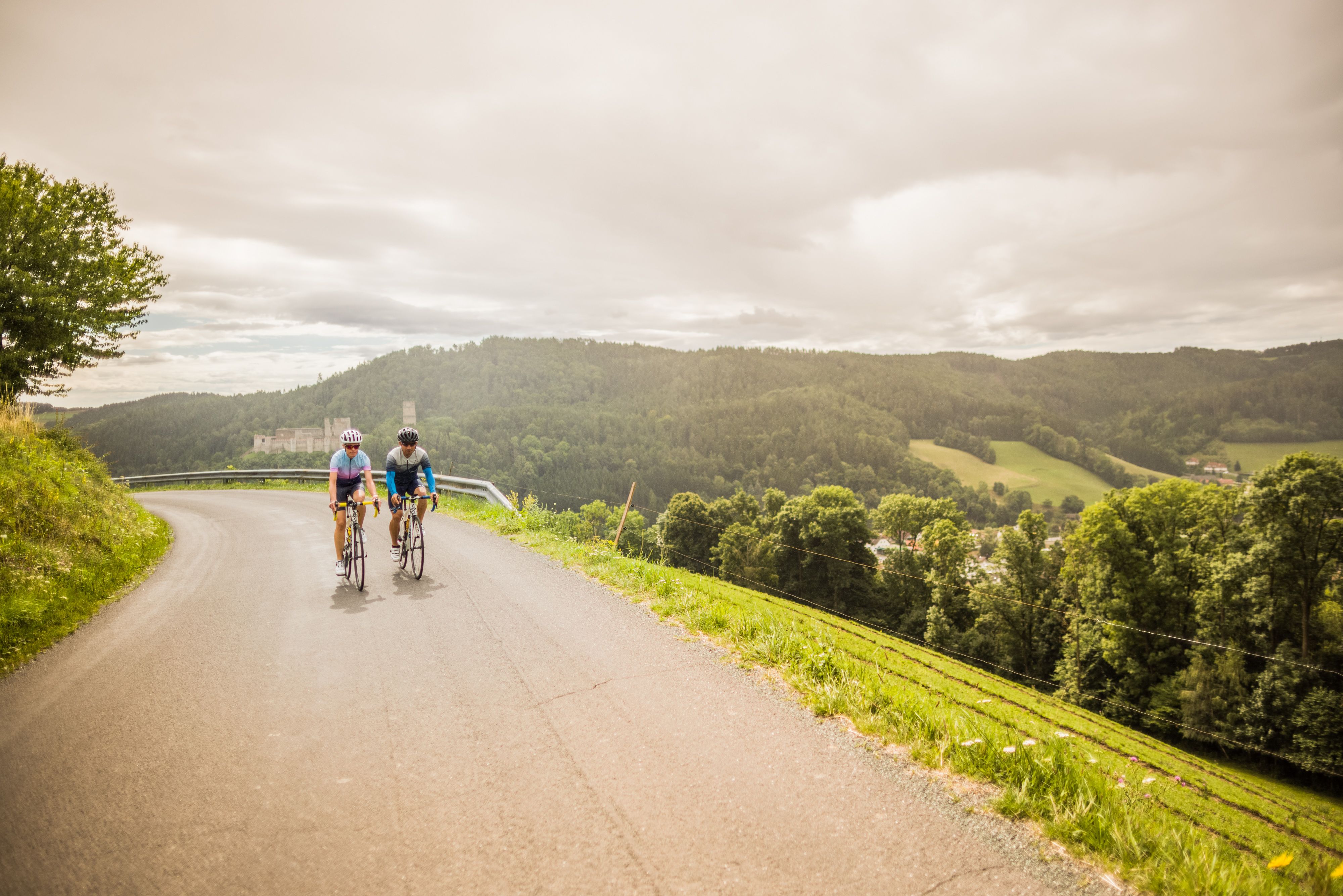 Dvaja ľudia na bicykloch na ceste v kopcovitej krajine s hradom v pozadí.