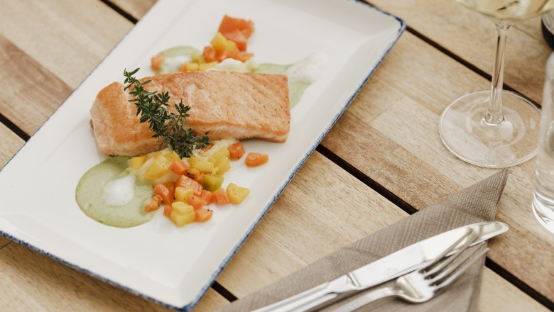 Na drevenom stole s pr&iacute;borom a poh&aacute;rom na v&iacute;no je tanier vypr&aacute;žan&eacute;ho lososa so zeleninou a zelenou om&aacute;čkou, ozdoben&yacute; vetvičkou byliniek.