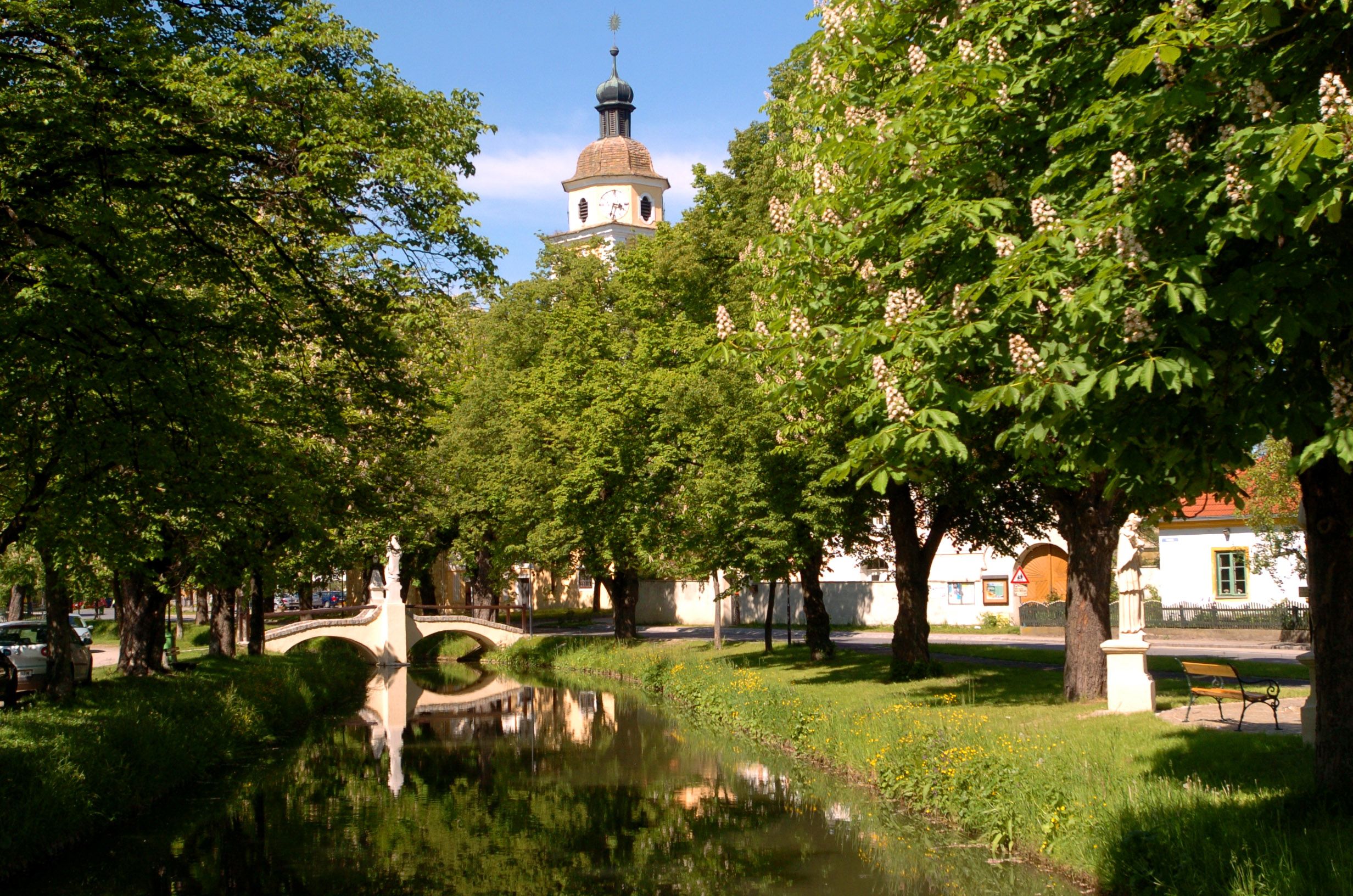 Idylický park s malou riekou, mostom a kostolnou vežou v pozadí.