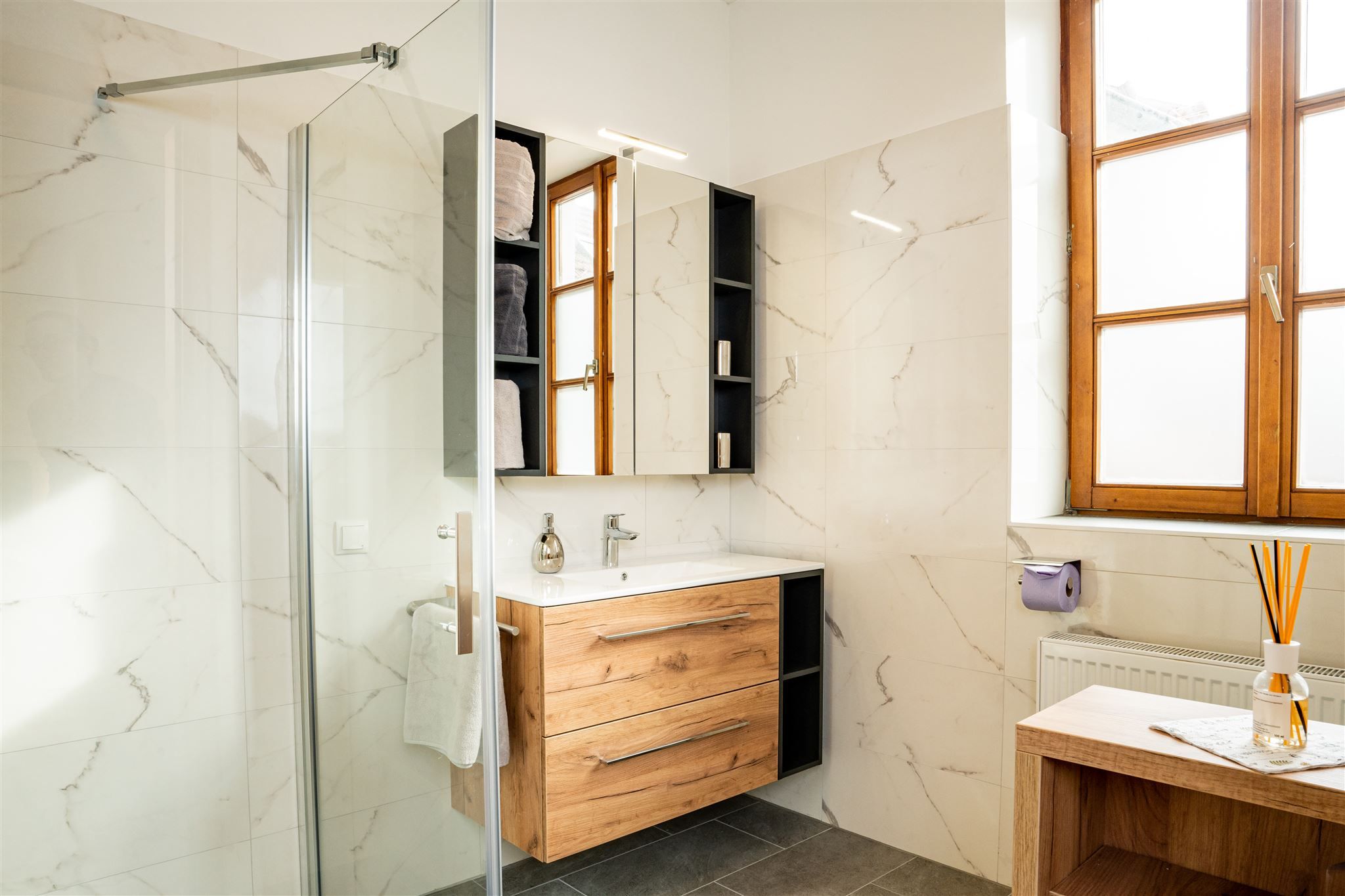 Moderná kúpeľňa s dreveným nábytkom, veľkým predným oknom a skleneným sprchovacím kútom.