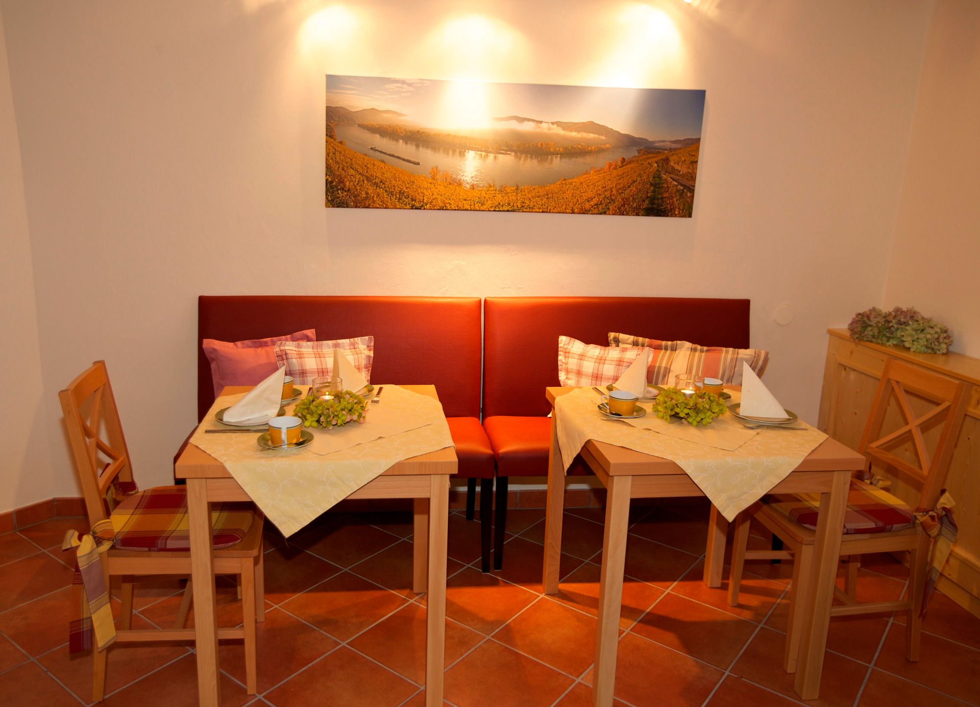 Útulná raňajková miestnosť s dvoma prestretými stolmi, červenými lavicami a krajinkou na stene.