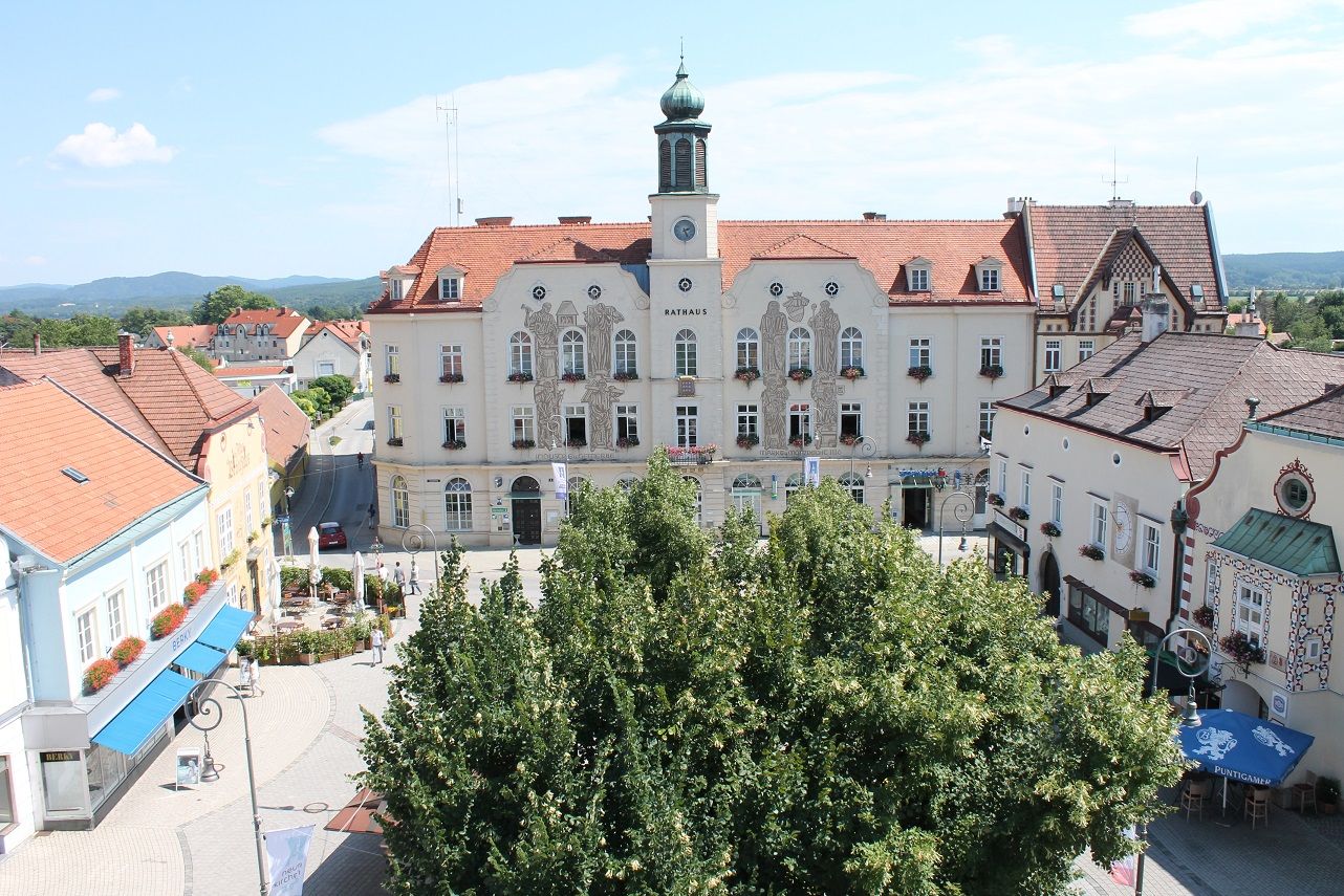 Historická radnica na slnečnom hlavnom námestí s okolitými budovami a stromami.