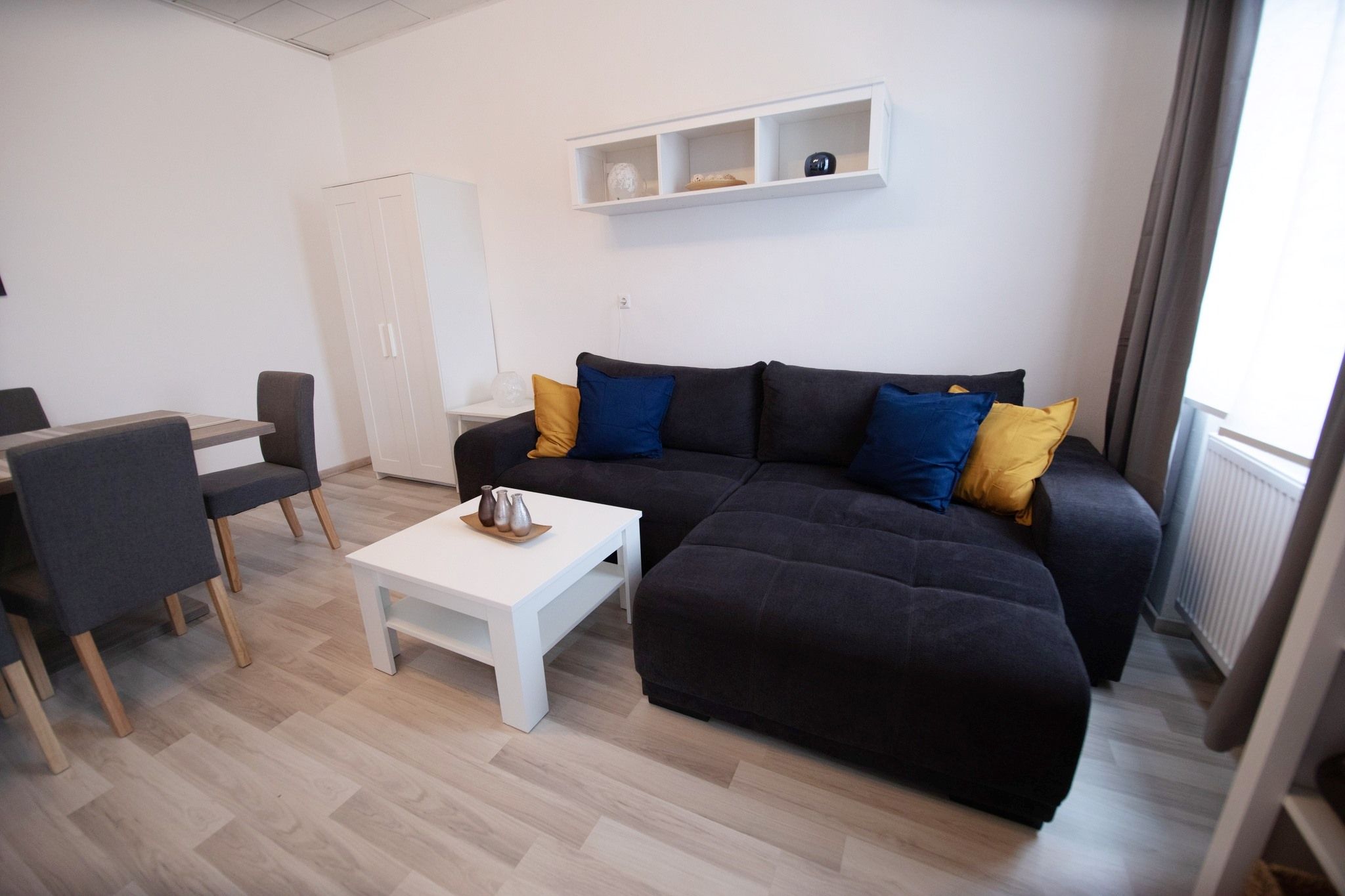 Moderný obývací priestor so sivou pohovkou, farebnými vankúšmi, bielym konferenčným stolíkom a jedálenským stolom so stoličkami.