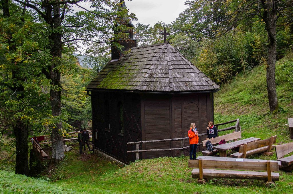 Malý drevený kostolík v lese s dvoma ľuďmi a drevenými lavicami v popredí.