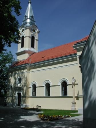 Farský kostol v Tattendorfe so žltou fasádou a červenou strechou, obklopený stromami.