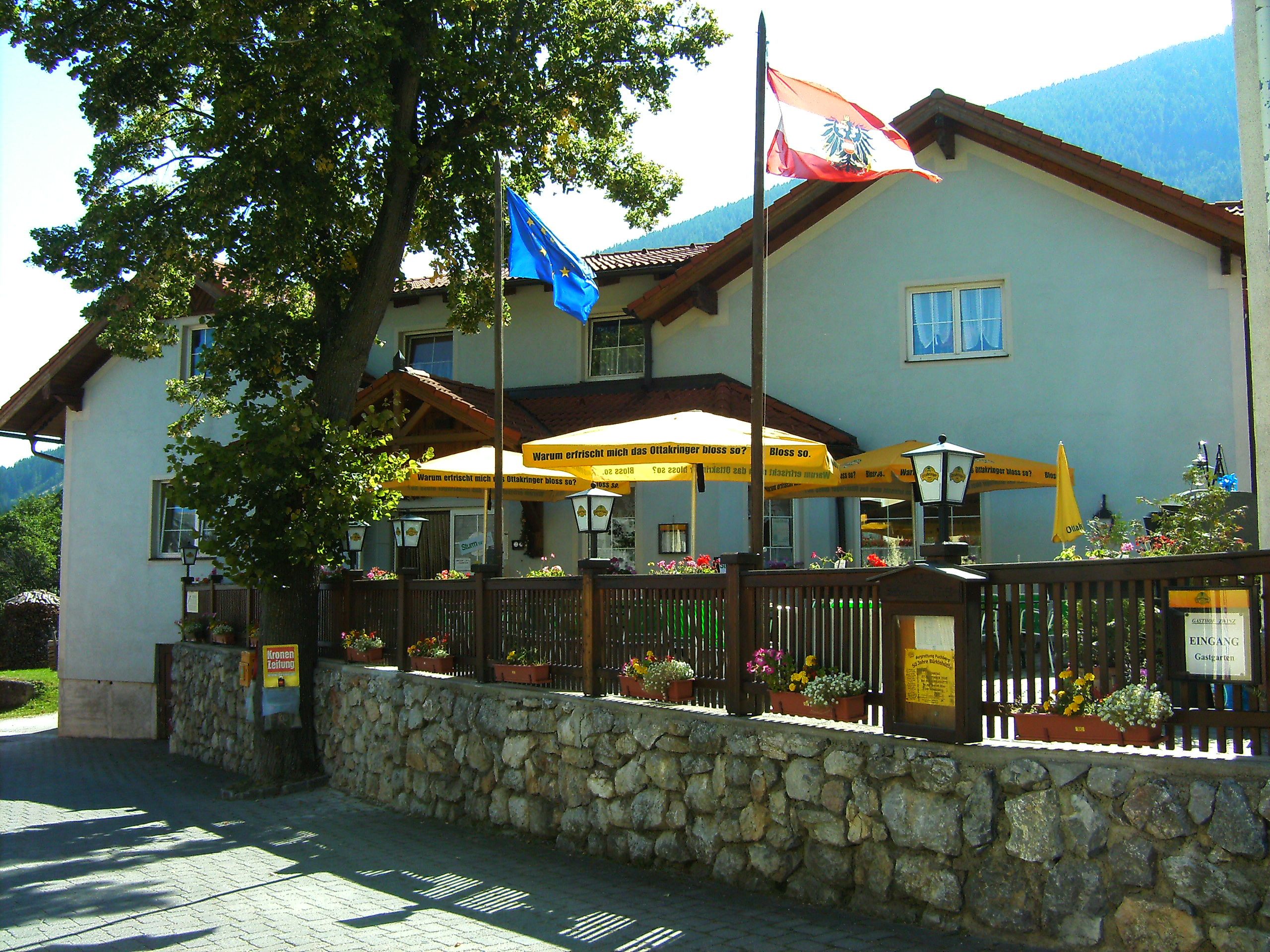 Gasthof Zwinz s terasou a slnečníkmi, obklopený kvetmi a vlajkami.