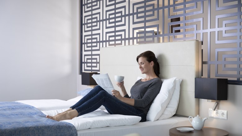 Žena odpoč&iacute;va na posteli v modernej hotelovej izbe, č&iacute;ta noviny a drž&iacute; &scaron;&aacute;lku. Dekorat&iacute;vna stena v pozad&iacute;.