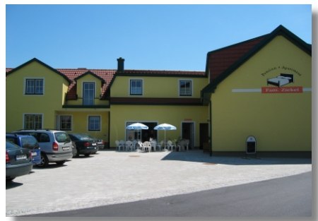 Žlt&aacute; budova s n&aacute;pisom "Pension Ziekel", pred ňou aut&aacute; a stoly so slnečn&iacute;kmi.