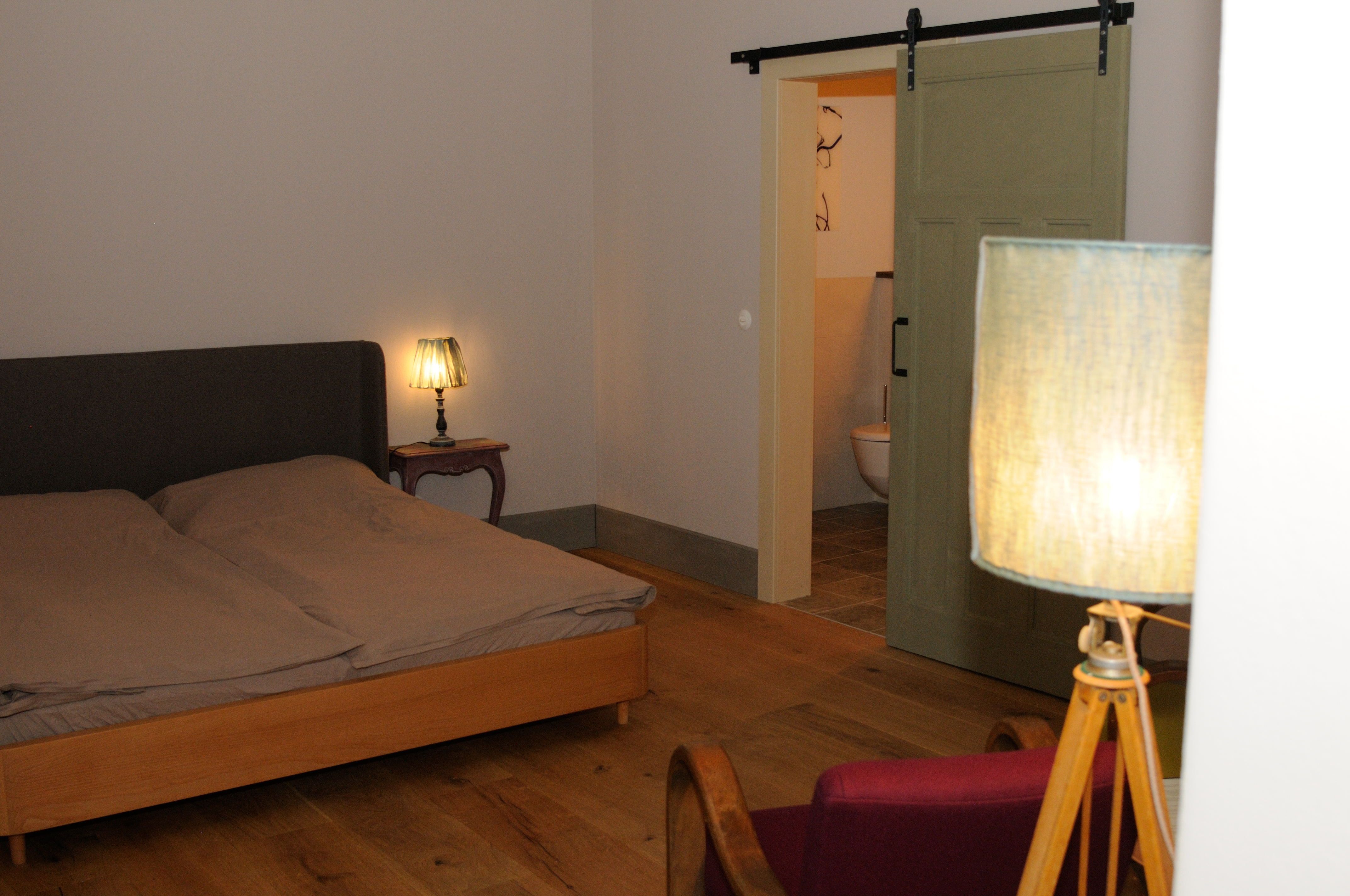 Útulná spálňa s manželskou posteľou, nočnou lampou a posuvnými dverami do kúpeľne.