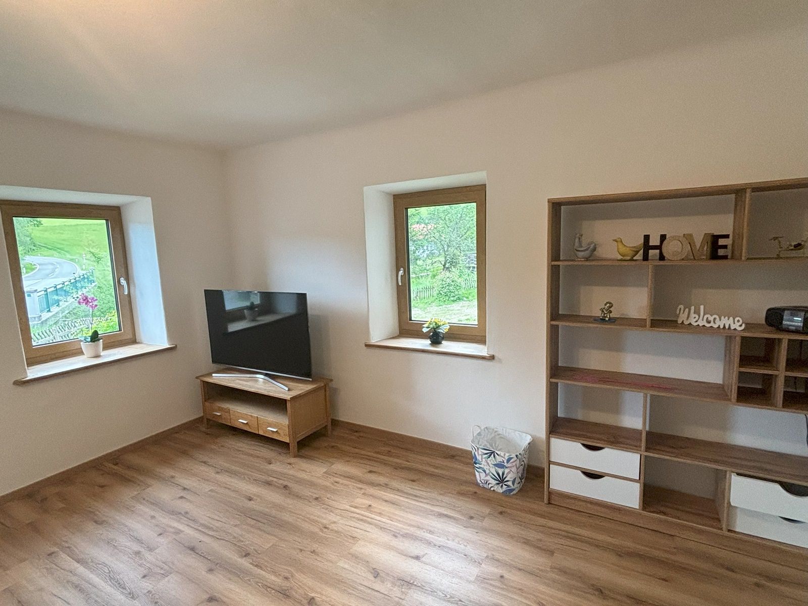 Obývacia izba s drevenou podlahou, televízorom na stole, dvoma oknami a policou s dekoráciami.