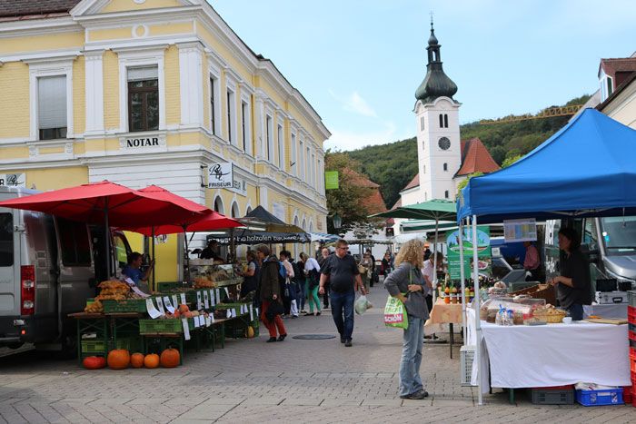 Živý farmársky trh so stánkami a návštevníkmi pred žltou budovou a kostolom v pozadí.