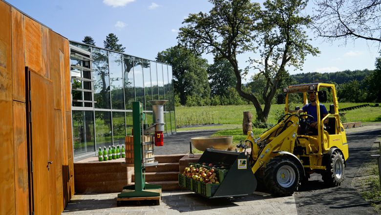 Žltý elektrický Hoftrac preváža debny s jablkami pred modernou budovou na farme.