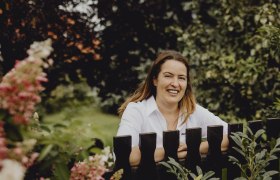 Žena v bielej košeli sa usmieva za dreveným plotom v záhrade.