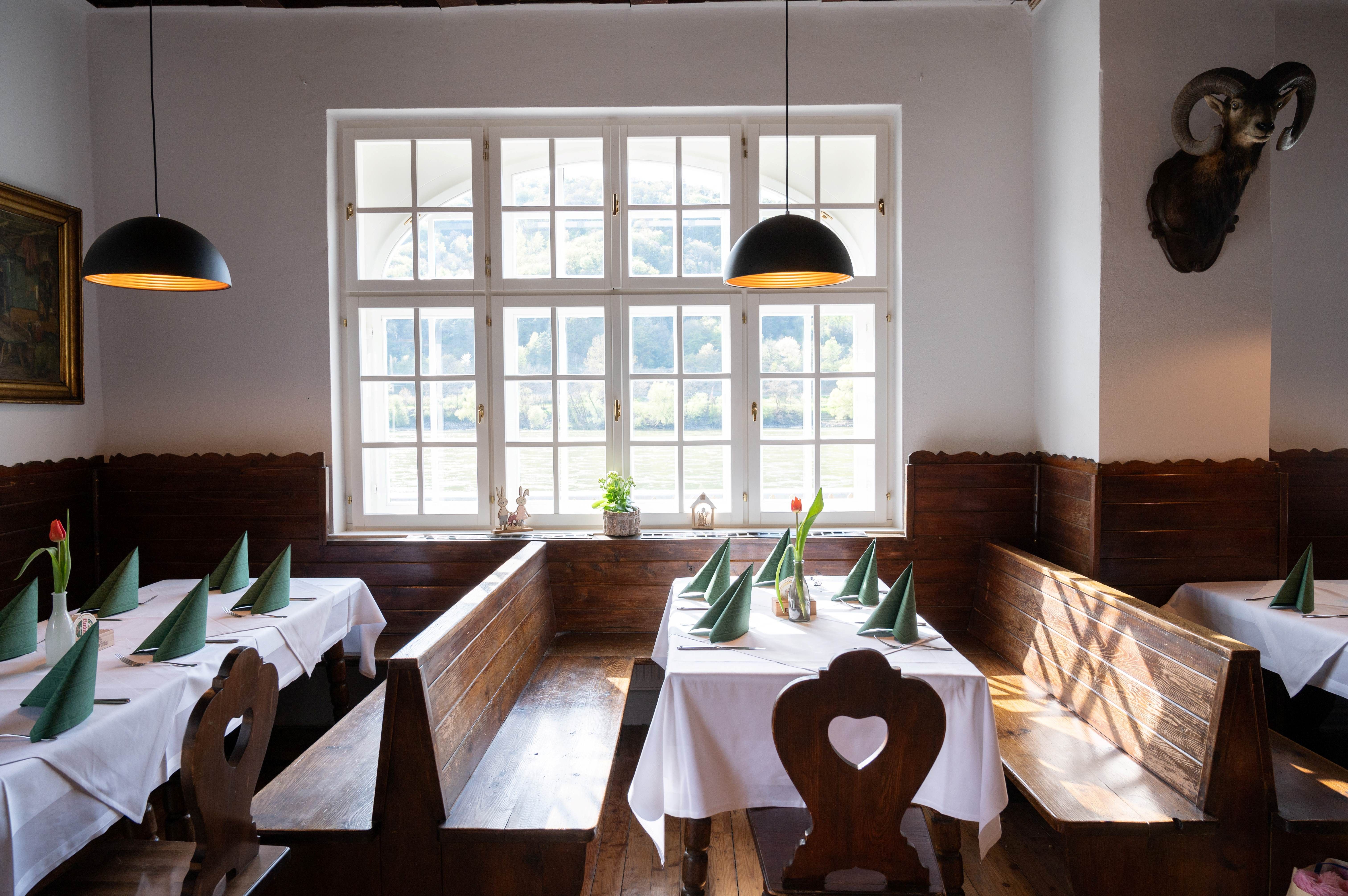 Útulná jedáleň s drevenými lavicami, bielymi obrusmi a zelenými obrúskami. Veľké okná s výhľadom na prírodu.