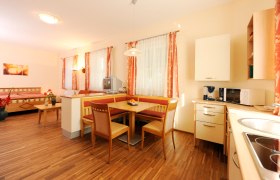 Svetl&aacute; izba s drevenou podlahou, jed&aacute;lensk&yacute;m stolom, kuchyňou a posteľou.