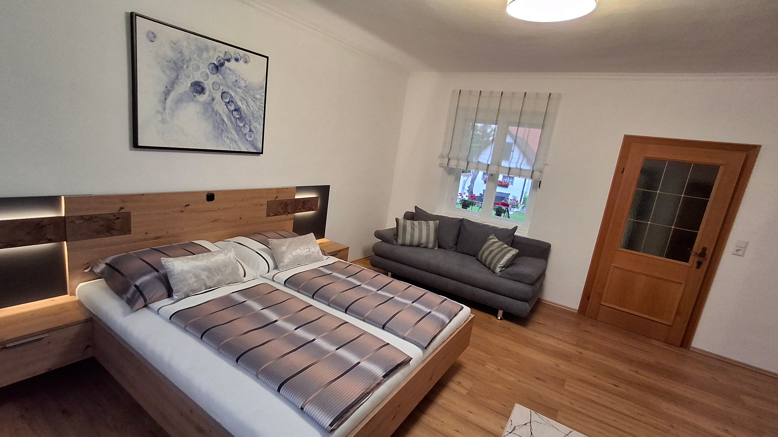 Moderná spálňa s manželskou posteľou, sivou pohovkou, drevenou podlahou a abstraktným umeleckým dielom na stene.