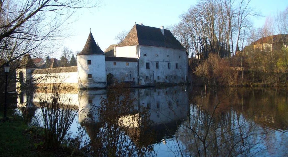Historická budova s vežami sa odráža v pokojnej vodnej ploche obklopenej holými stromami.