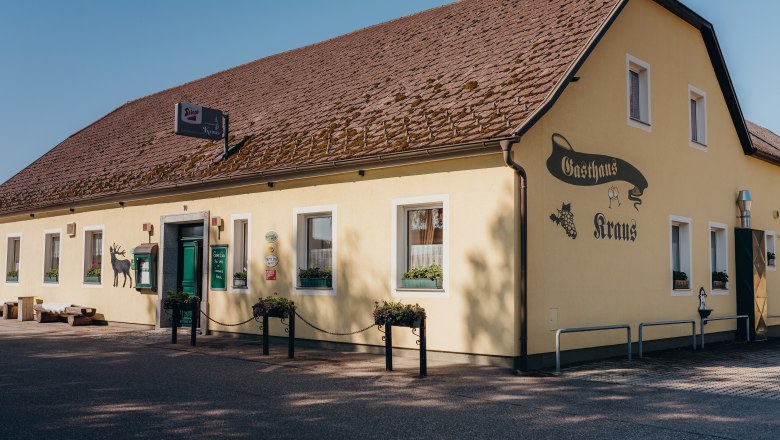 Žltý hostinec s hnedou strechou a zelenými dverami s nápisom "Gasthaus Kraus".
