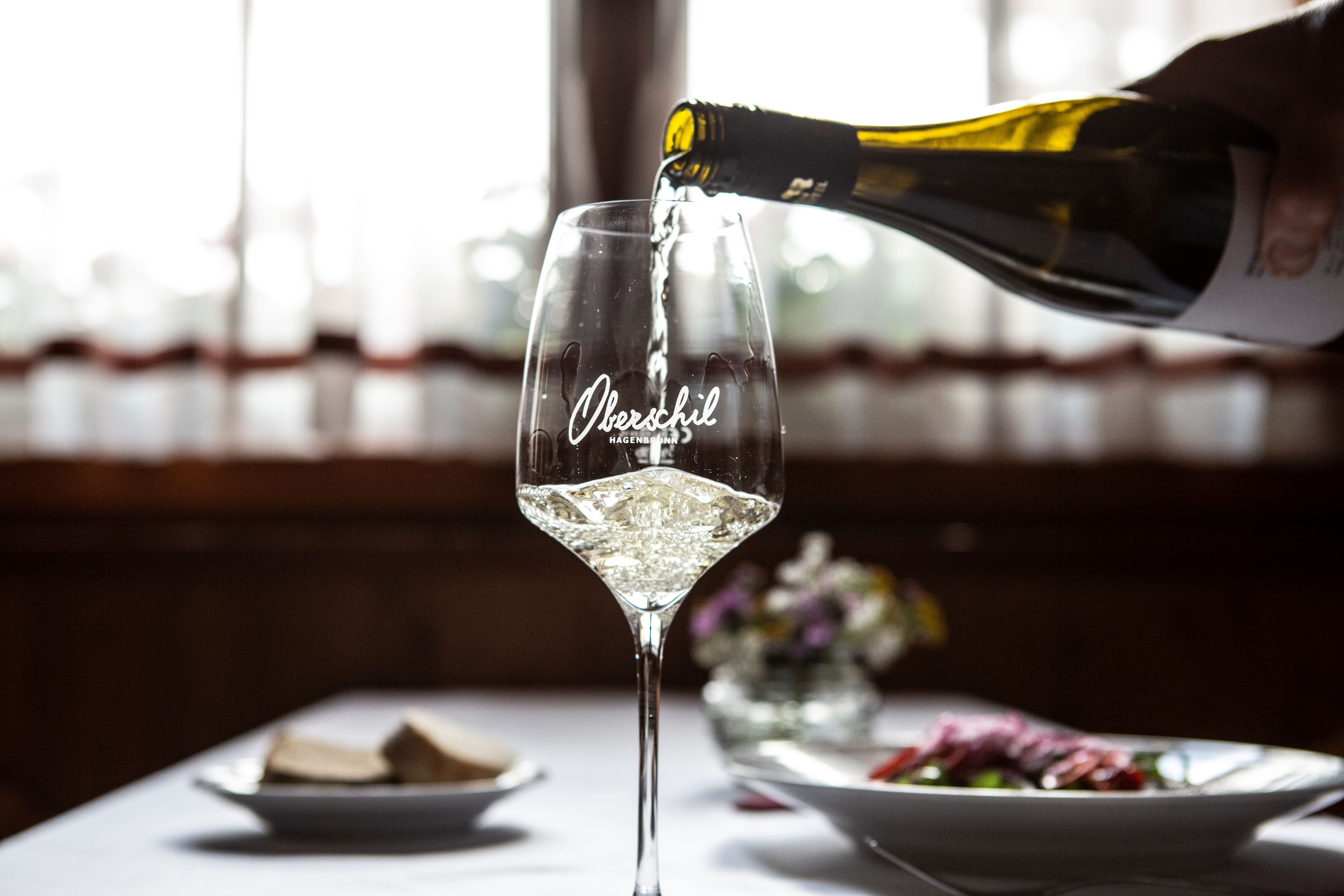 Nalievajú pohár bieleho vína, v pozadí je prestretý stôl s kvetmi.