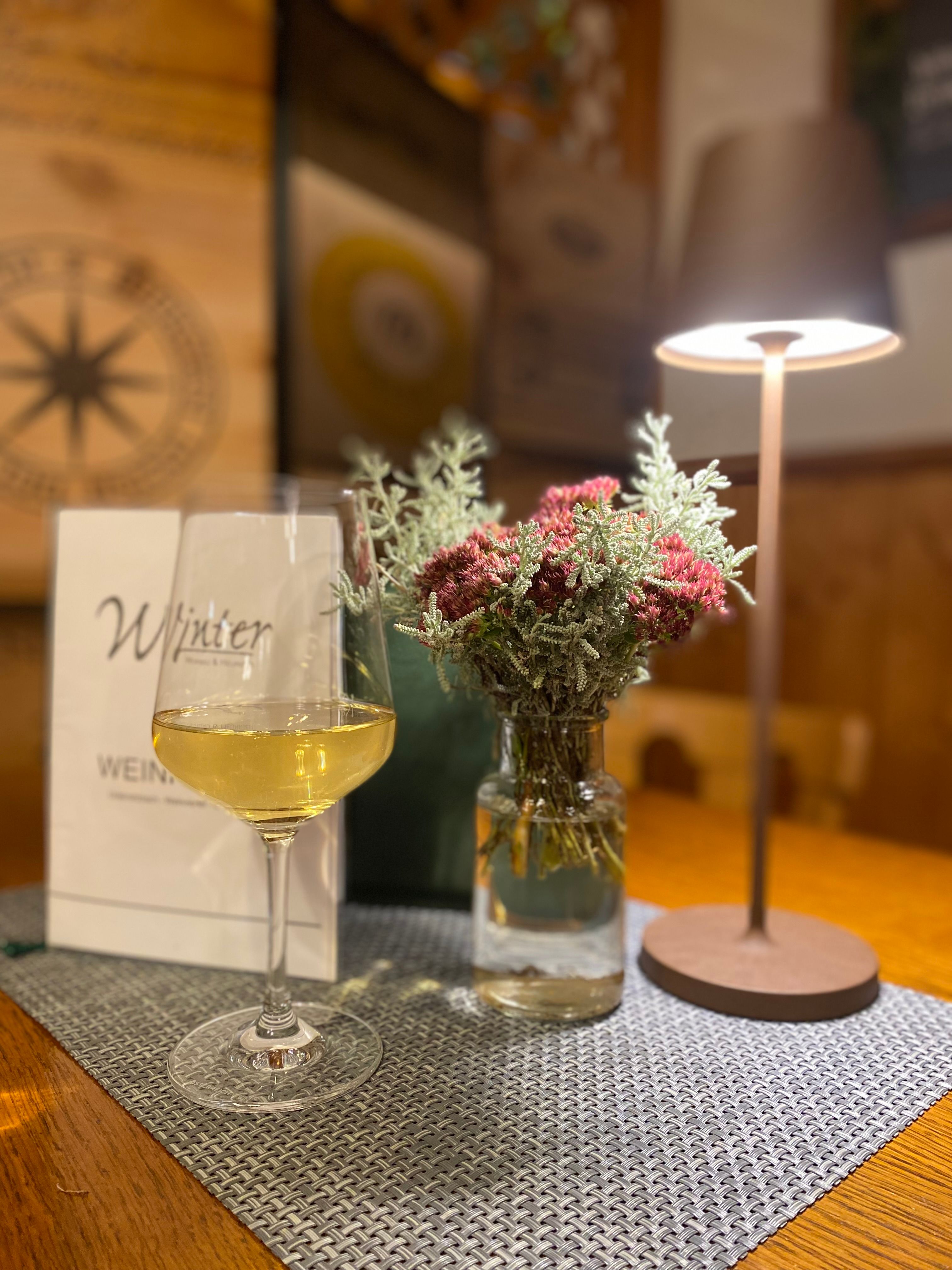 Pohár s bielym vínom stojí na stole vedľa vázy s kvetmi a stolnej lampy.