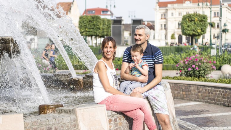 Eine Familie sitzt an einem Brunnen in einer Stadt mit historischen Gebäuden im Hintergrund.