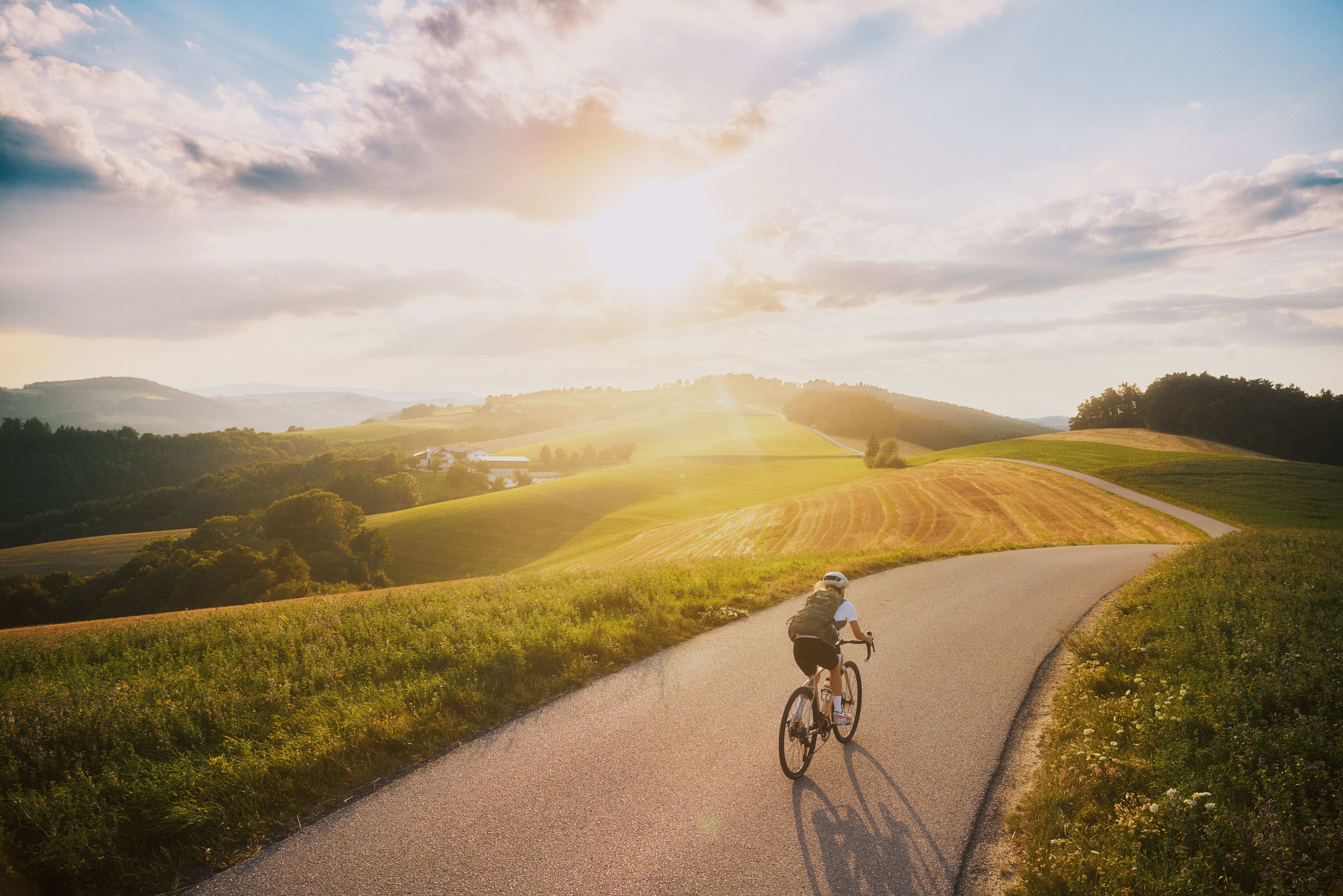 Cyklista jazdí po kľukatej vidieckej ceste kopcovitou krajinou pri západe slnka.