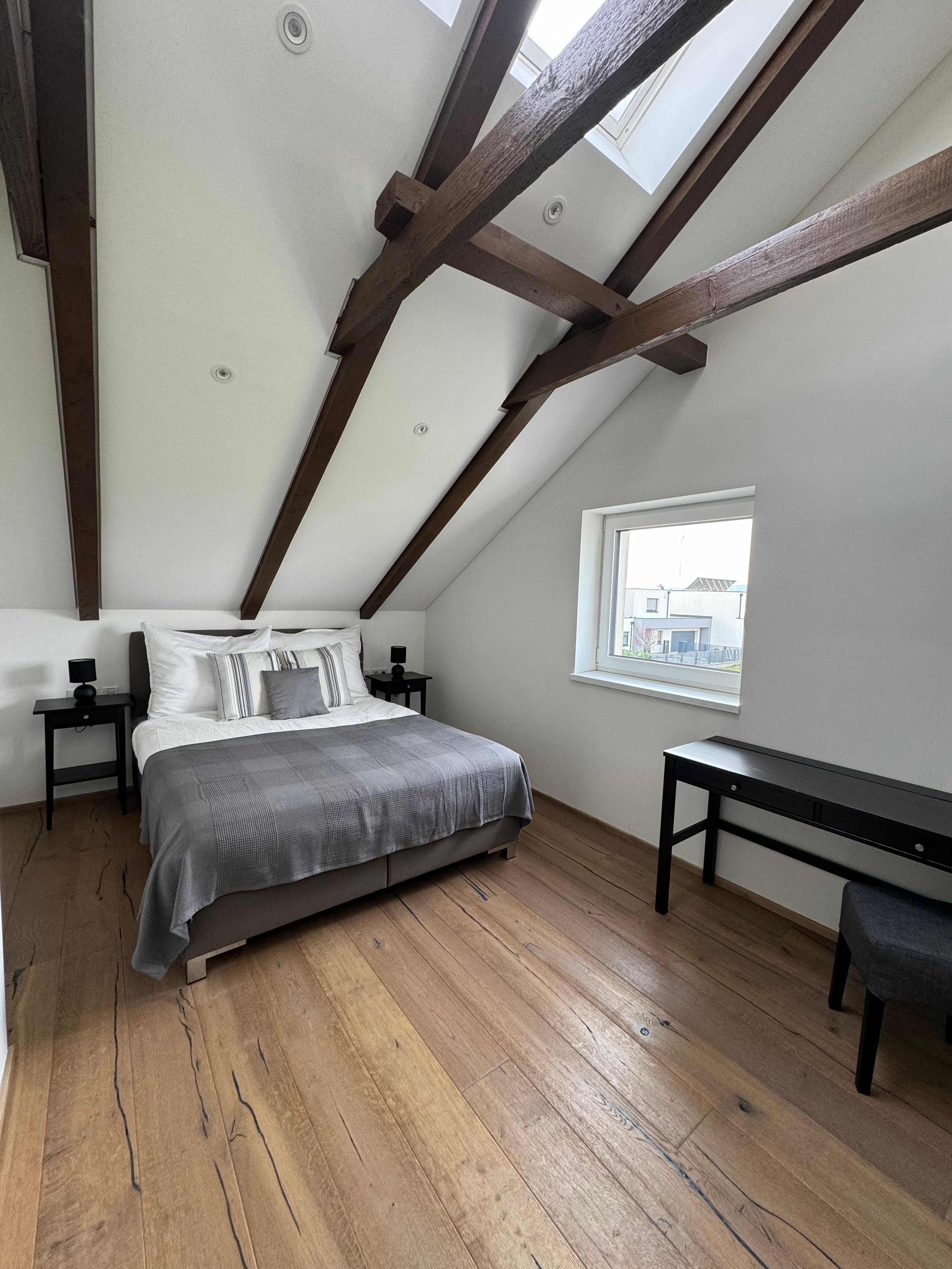 Moderná spálňa s dreveným trámovým stropom, manželskou posteľou, drevenou podlahou a oknom.