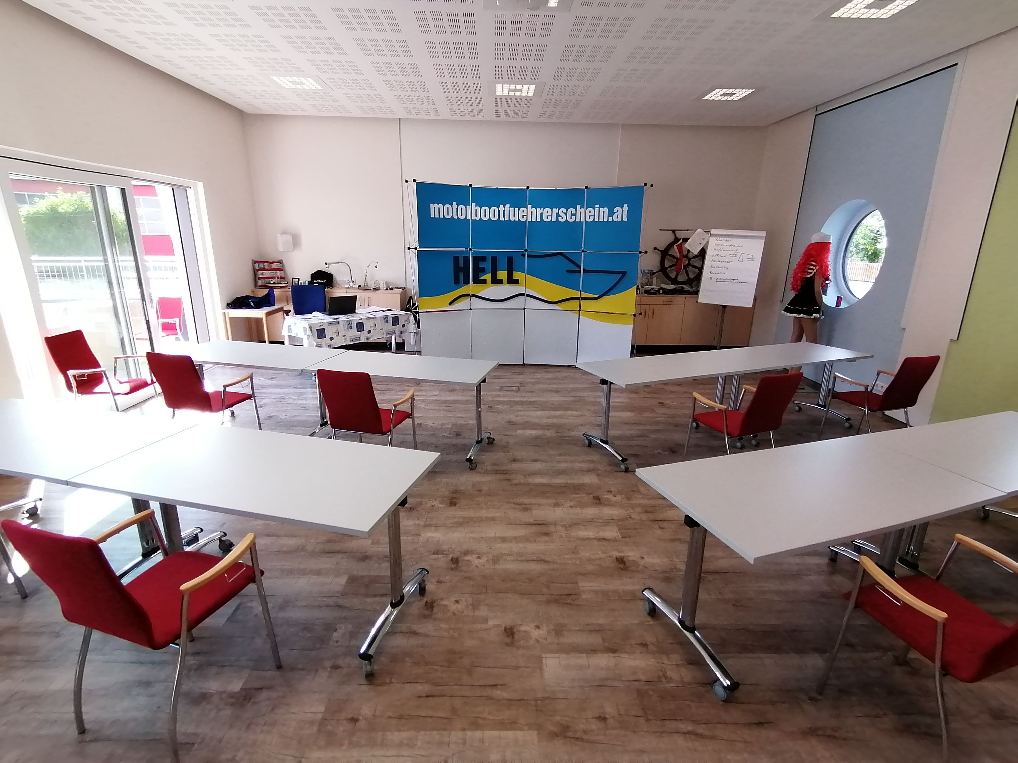 Školiaca miestnosť so stolmi a stoličkami, plagátom školy kapitána pekla, flipchartom a figurínou.