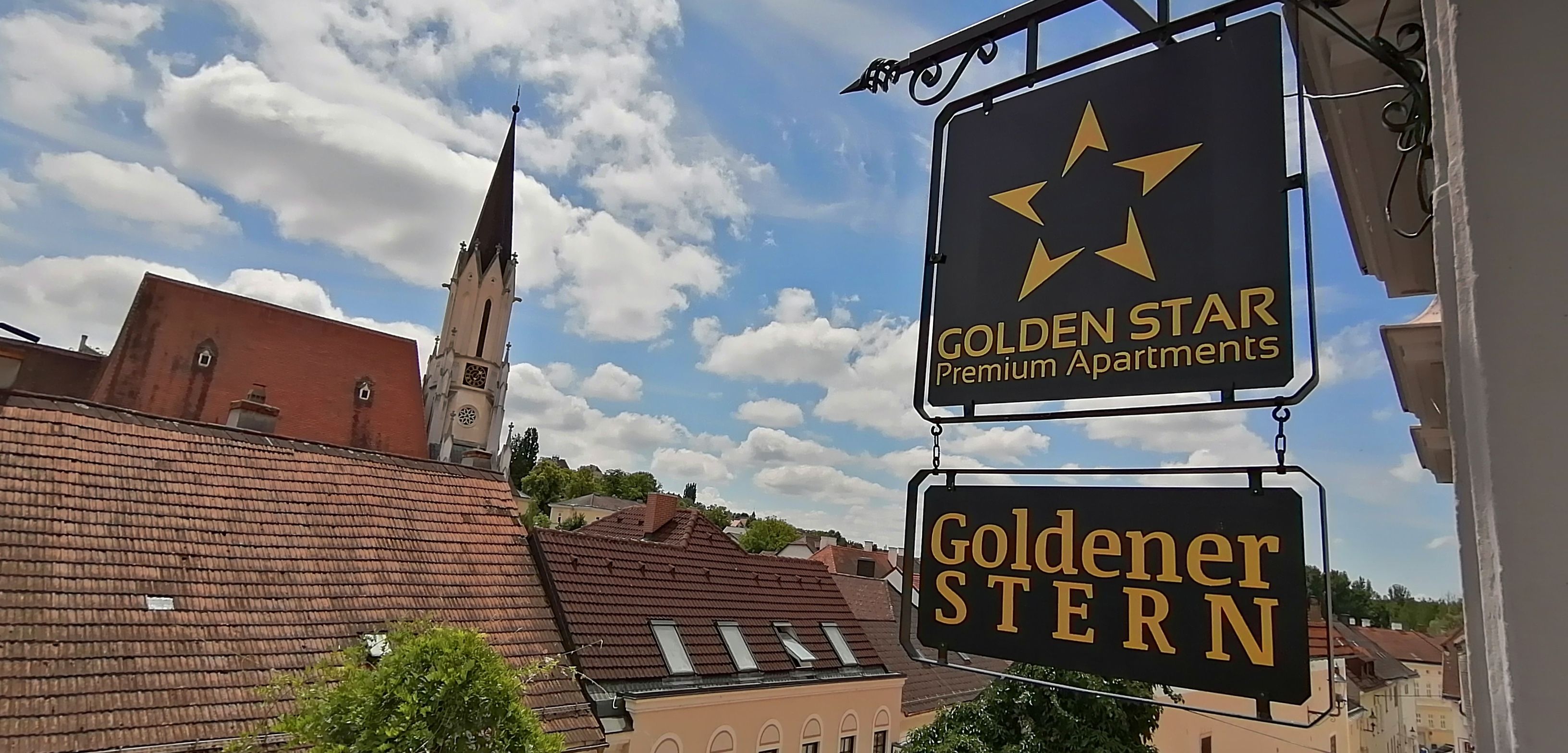 Pohľad na nápis "Golden Star Premium Apartments" a "Goldener Stern" pred kostolnou vežou a strechami.