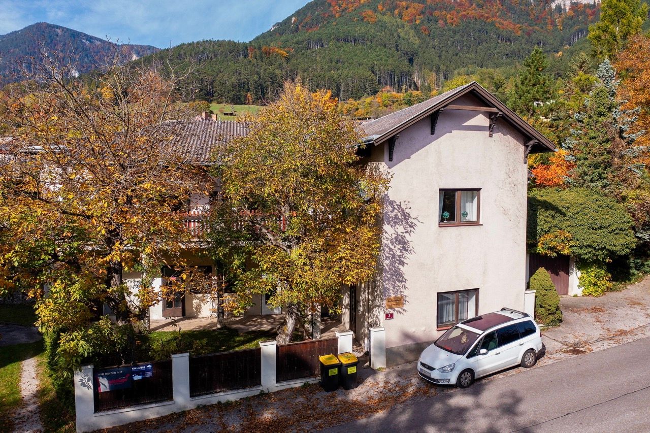 Dvojposchodová budova s jesennými stromami a zaparkovaným autom pred ňou, obklopená zalesnenými kopcami.