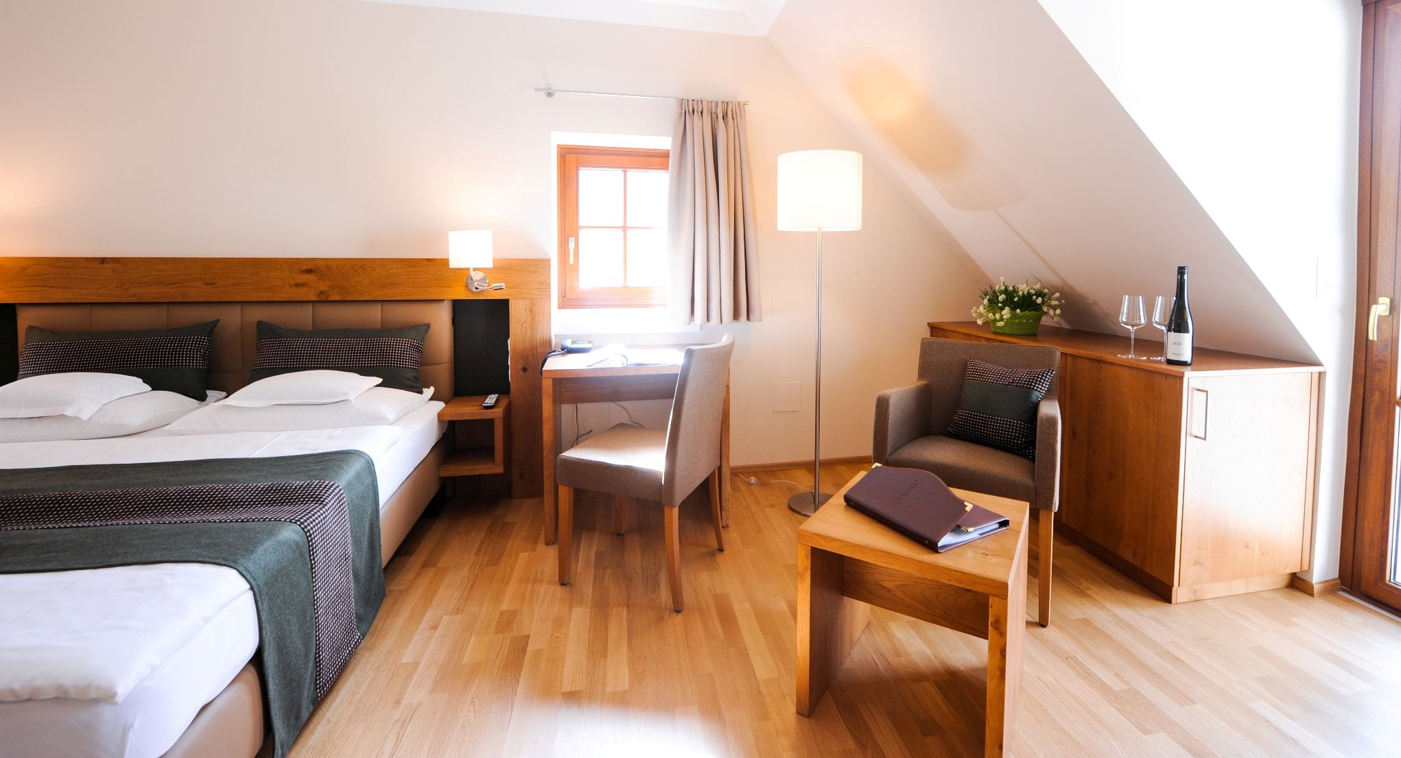 Útulná hotelová izba s manželskou posteľou, písacím stolom, kreslom a fľašou vína na skrini.