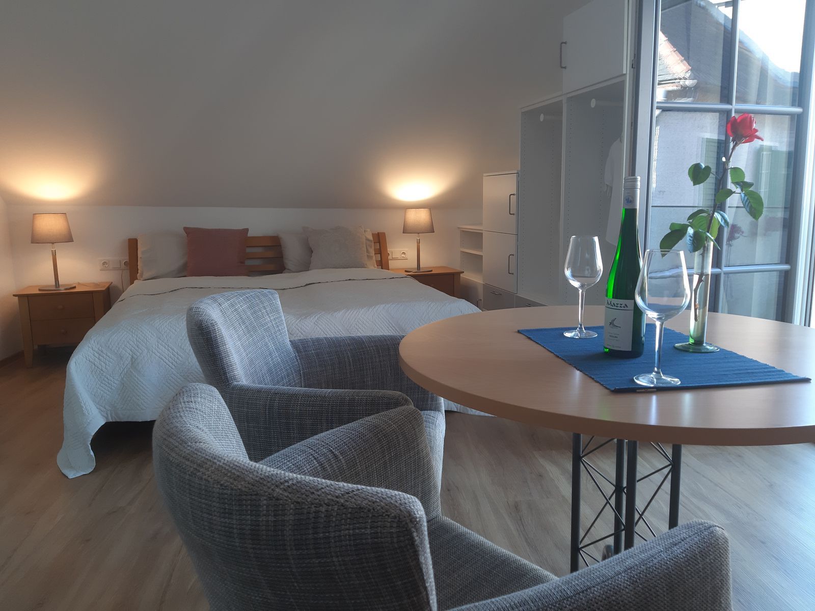 Útulná spálňa s manželskou posteľou, stolom, dvoma stoličkami, fľašou vína a pohármi.