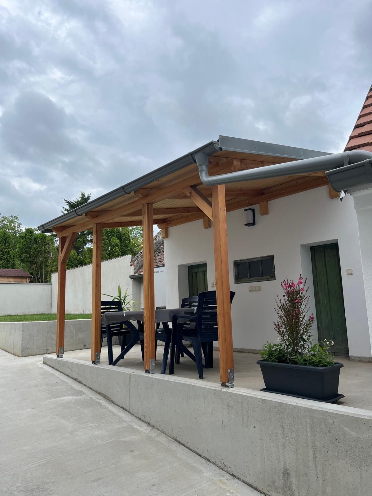 Krytá terasa so stolom a stoličkami, obklopená rastlinami.