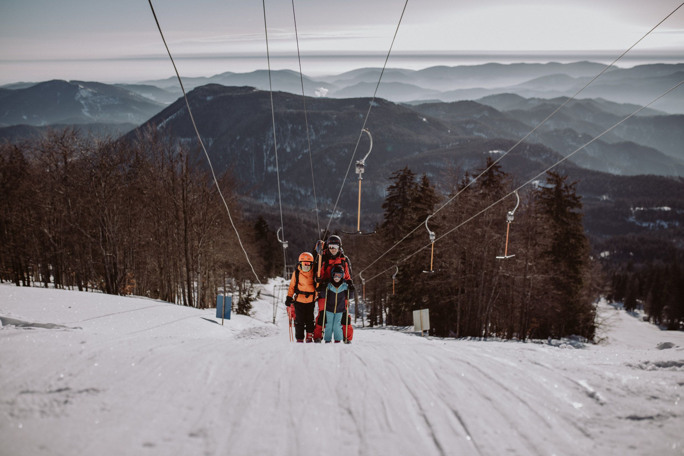 Traja lyžiari na vleku v lyžiarskej oblasti Unterberg, obklopení zasneženými horami a stromami.