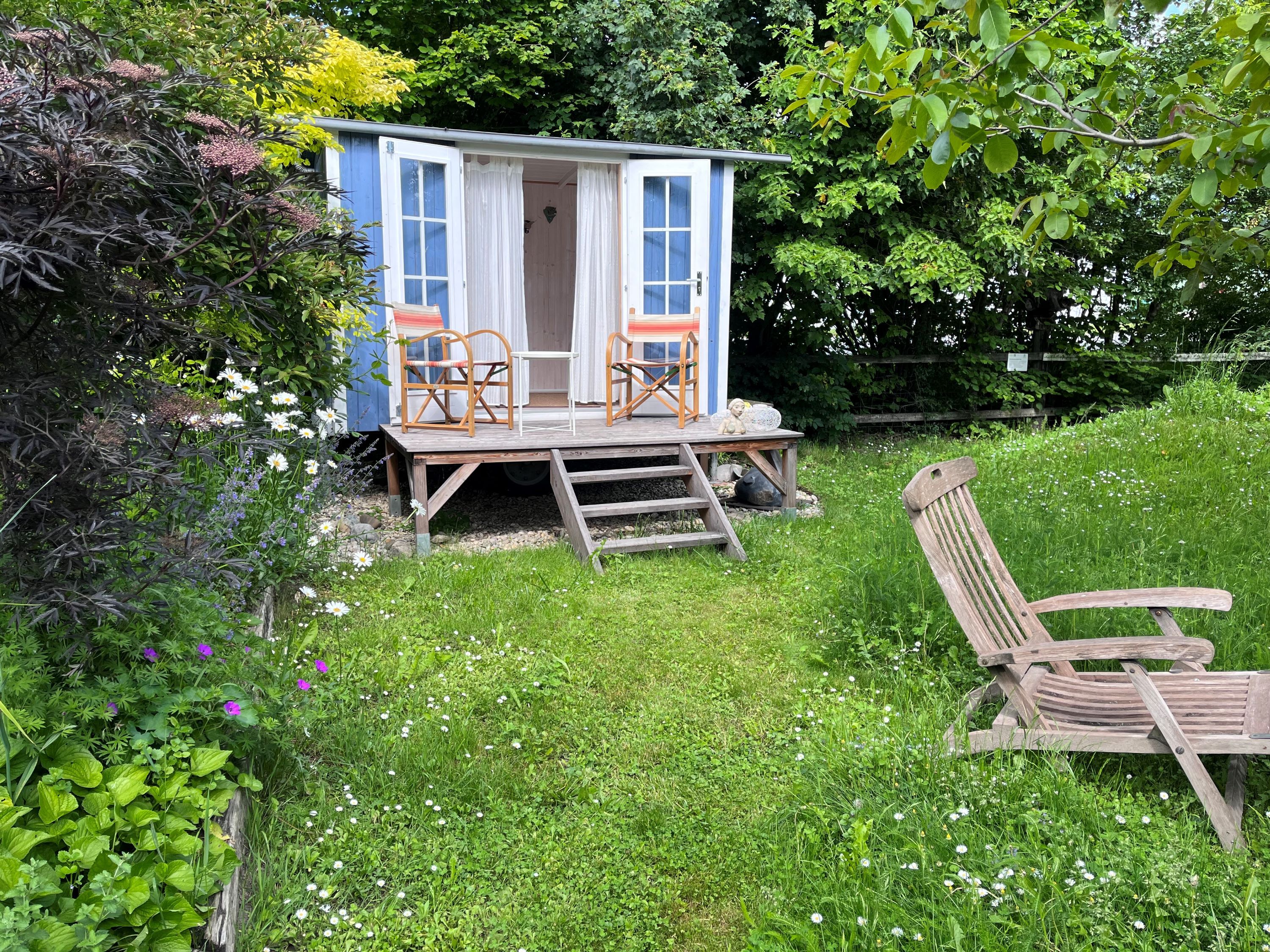 Modrý záhradný pavilón s dvoma stoličkami na drevenej verande, obklopený zelenou lúkou a farebnými kvetmi.