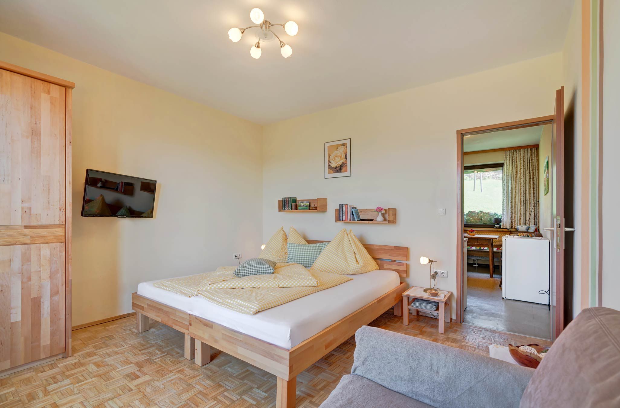 Útulná spálňa s drevenou podlahou, manželskou posteľou so žltou posteľnou bielizňou, TV s plochou obrazovkou na stene a otvorenou chodbou do kuchyne.