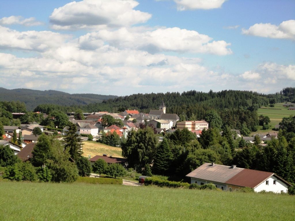 Panoráma trhového mestečka Martinsberg s kostolom a okolitými domami, obklopeného lesmi a poľami pod modrou oblohou.