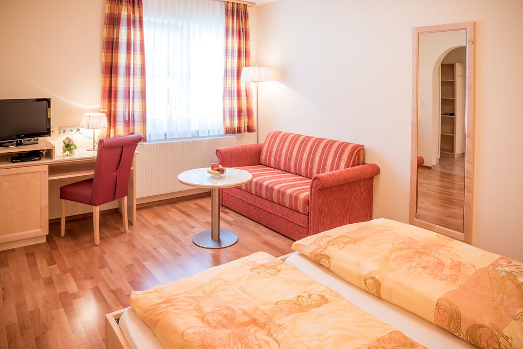 Útulná izba pre hostí s manželskou posteľou, červenou pohovkou, písacím stolom, TV a veľkým zrkadlom.
