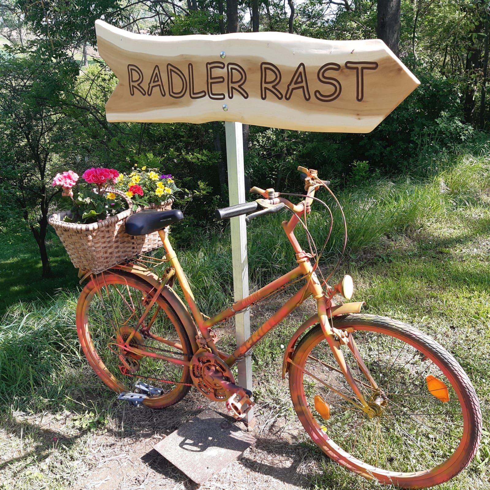 V zelenom prostredí stojí oranžový bicykel s košíkom na kvety vedľa drevenej tabule s nápisom „Radler Rast“.