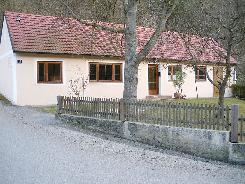 Jednopodlažný rekreačný dom s červenou škridlovou strechou a svetlým náterom, obklopený dreveným plotom a stromami.