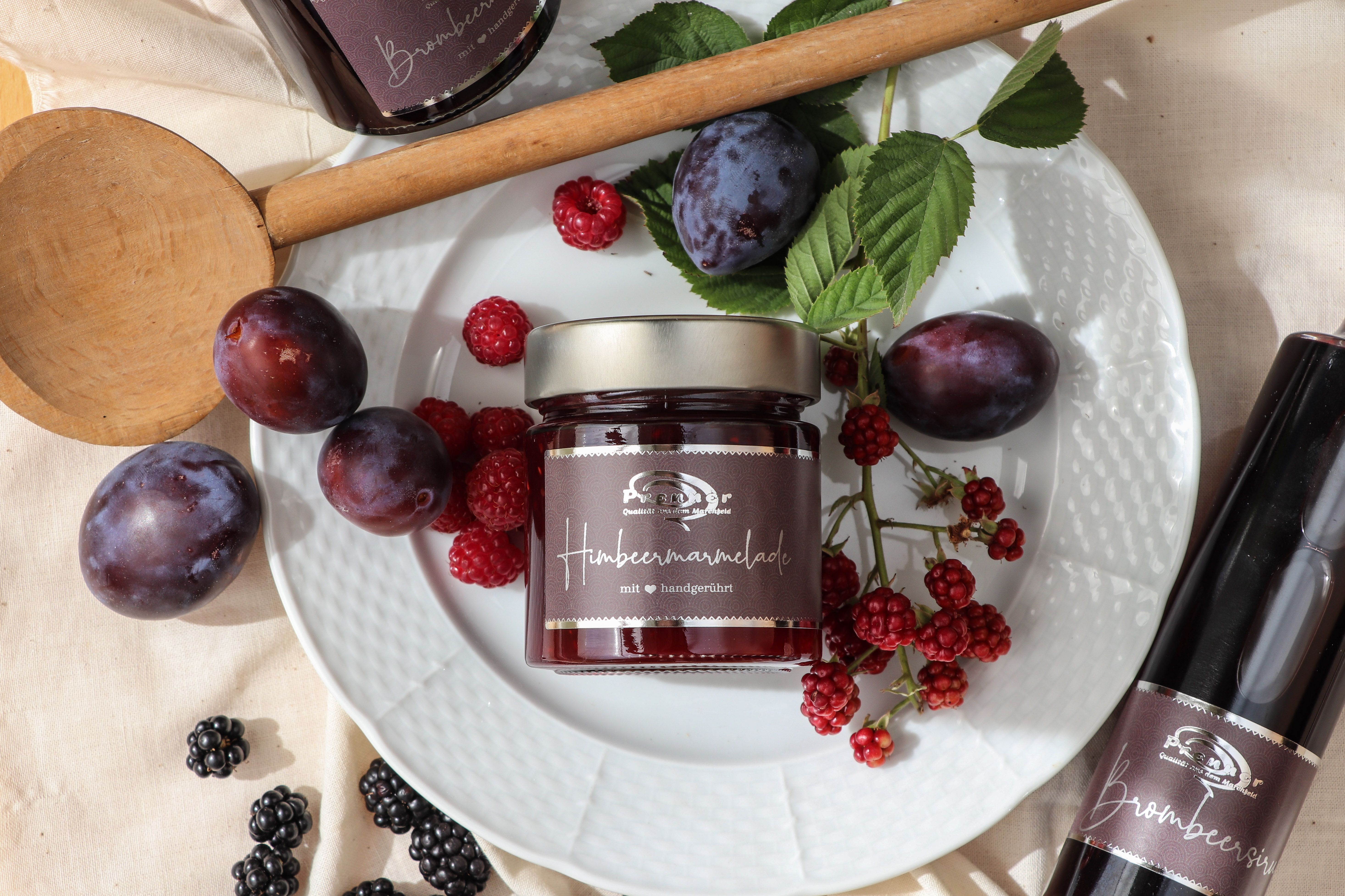 Pohár malinového džemu na tanieri s čerstvými slivkami, malinami a černicami, obklopený drevenou lyžicou a fľašou ostružinového sirupu.