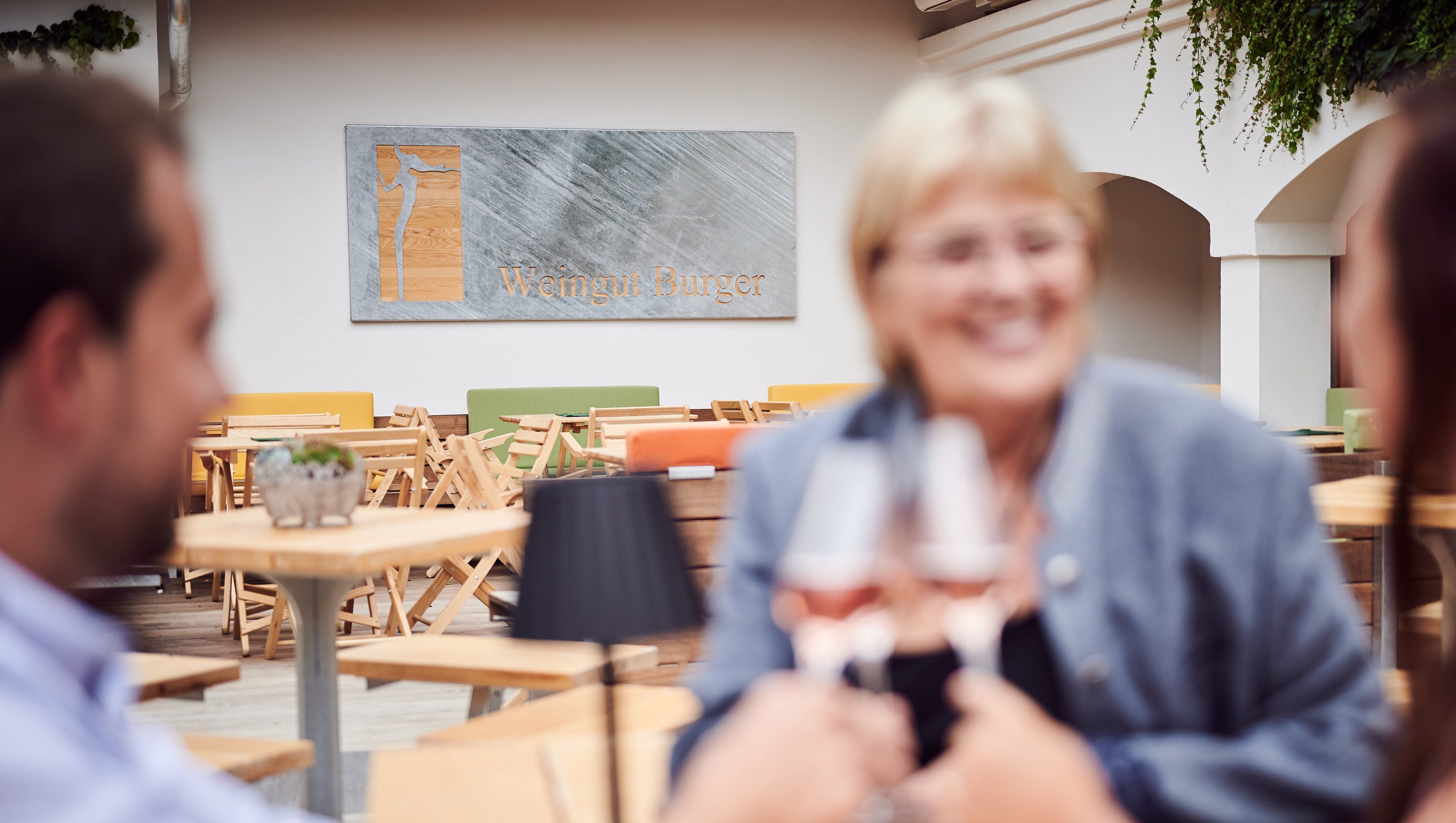 Traja ľudia si na nádvorí pripíjajú pohármi vína a v pozadí je nápis "Weingut Burger".